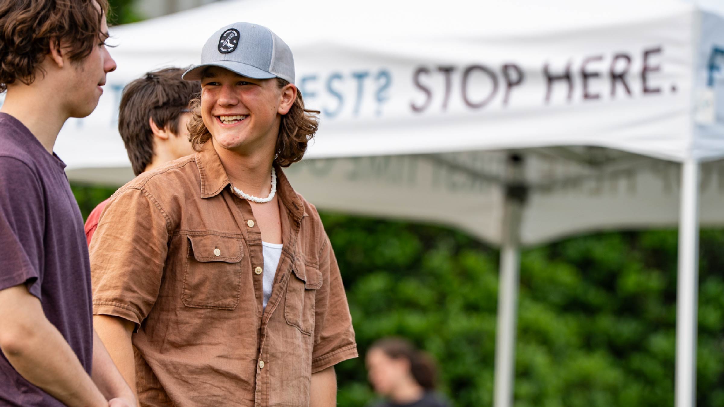A smiling teenage boy in a brown shirt and gray cap stands outdoors, chatting with friends by a white tent that partially reads “STOP HERE.” Greenery fills the background, capturing a candid moment perfect for photography & videography services.