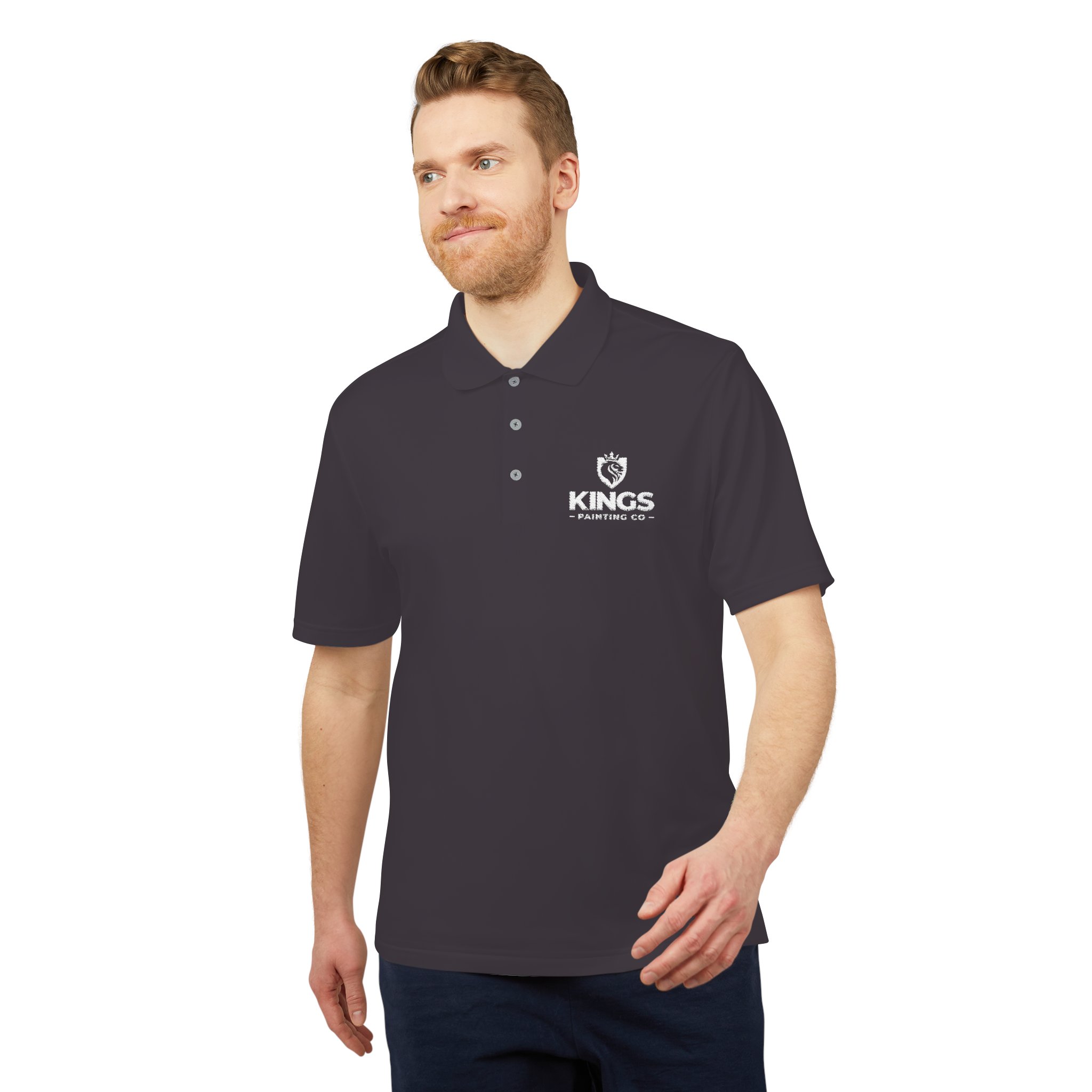 A man with light hair and a beard is standing and smiling, his right arm slightly bent, wearing a dark short-sleeve Kings Painting Co. Performance Polo by Under Armour® featuring a white logo on the left chest.